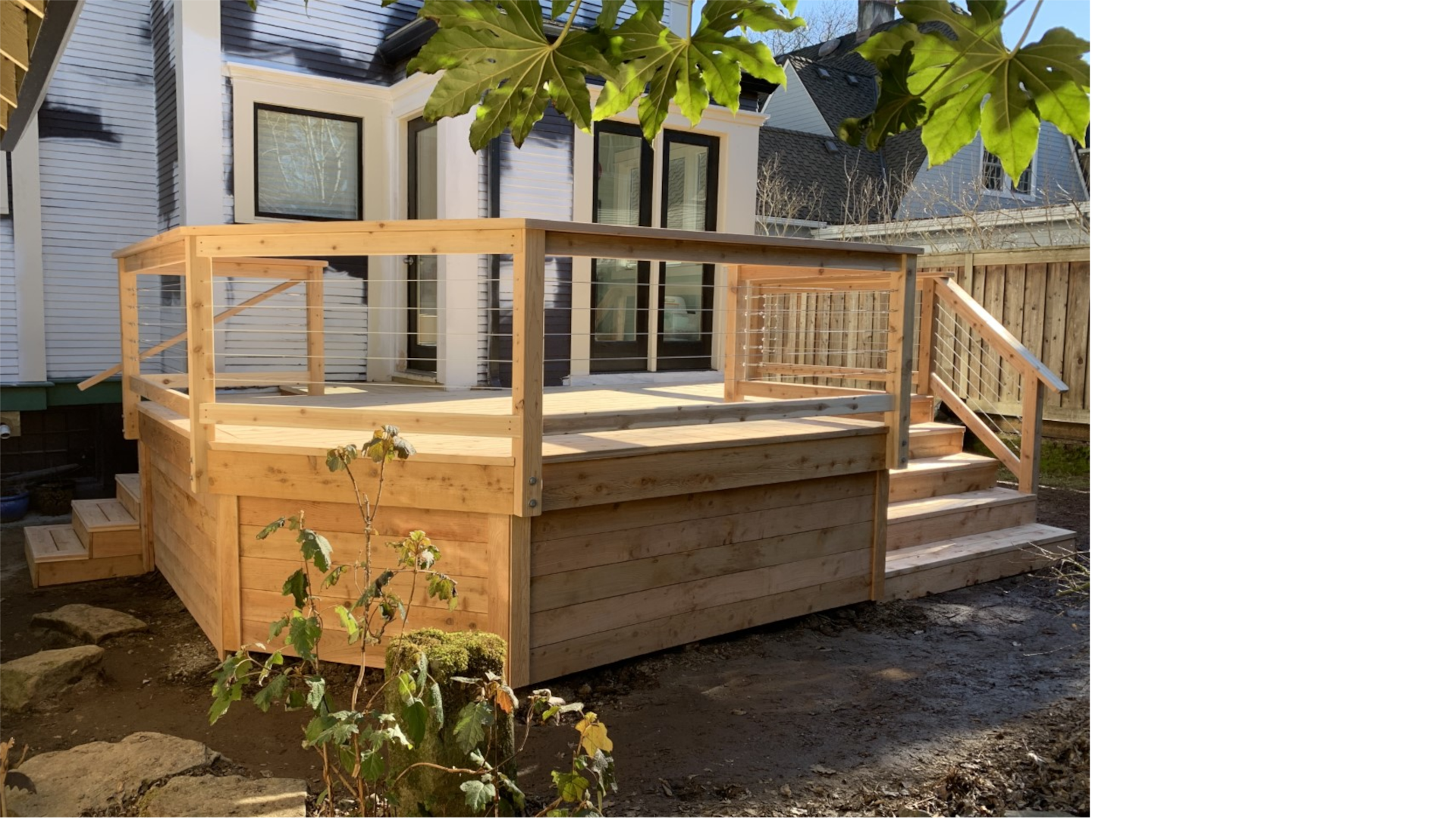 Elevated cedar deck with horizontal stainless-steel cable railing, attached to a white century home, shown on a sunny day.
